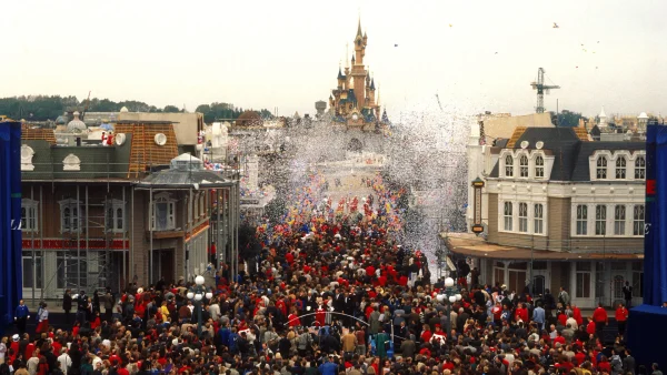 Une foule nombreuse envahit une rue de Disneyland Paris, tandis que des confettis tombent devant le Château de la Belle au Bois Dormant.