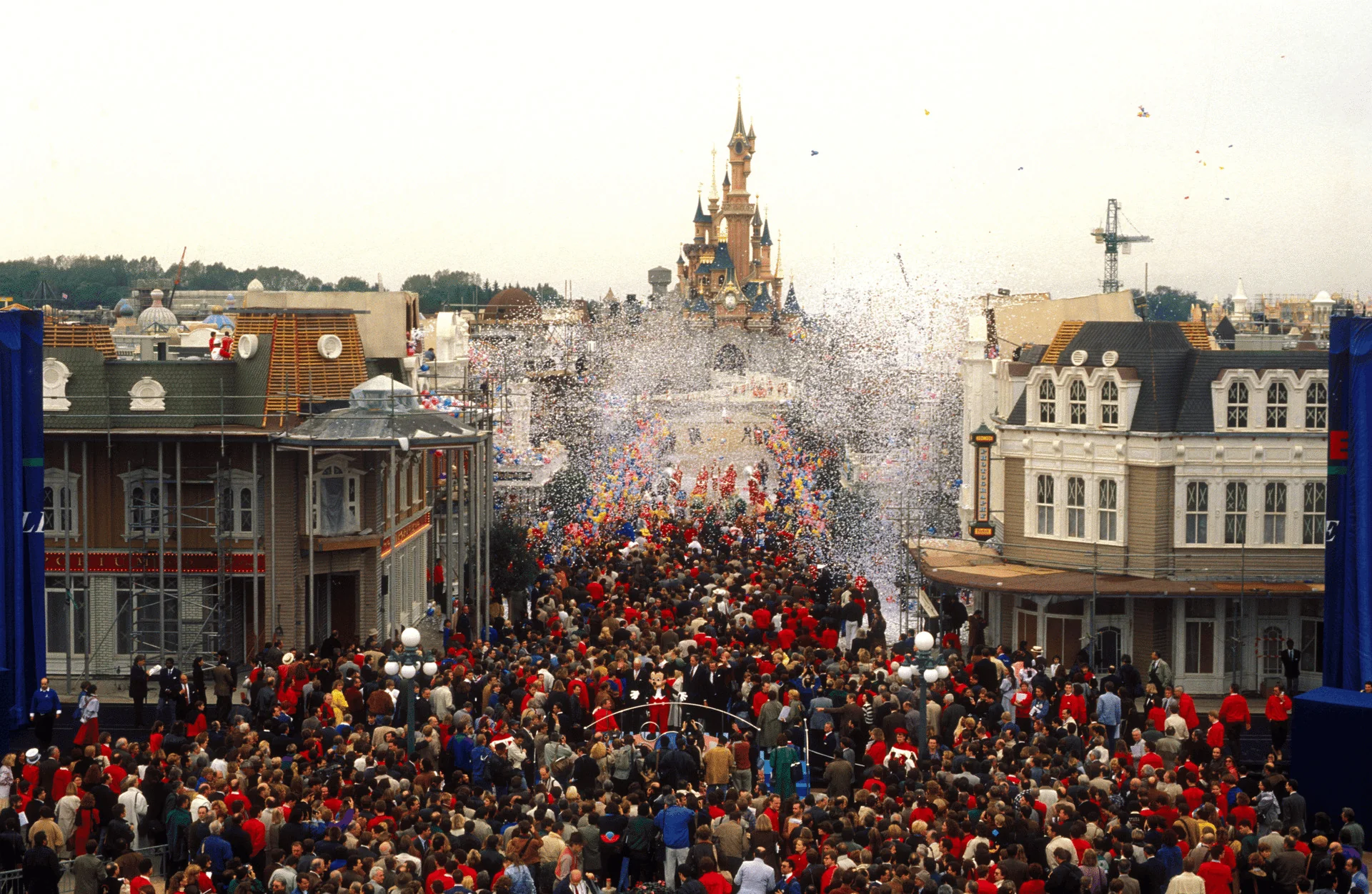 Une foule nombreuse envahit une rue de Disneyland Paris, tandis que des confettis tombent devant le Château de la Belle au Bois Dormant.