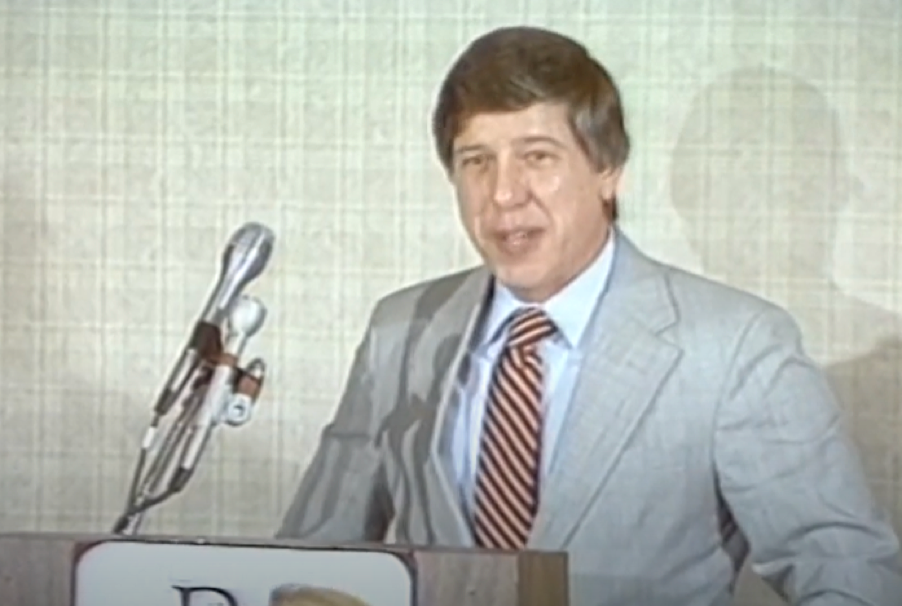 Un homme vêtu d'un costume clair et d'une cravate rayée se tient debout derrière un pupitre équipé de microphones, s'exprimant devant un fond neutre, peut-être pour annoncer la création de Disney Channel le 18 avril 1983.