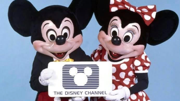 Deux personnes déguisées en Mickey et Minnie Mouse tiennent une pancarte arborant le logo de Disney Channel, célébrant la création de la chaîne le 18 avril 1983, sur un fond bleu uni.