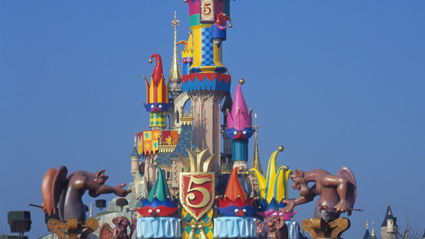 Entrée colorée du château à Disneyland Paris, décorée de grands personnages de dessins animés et d'un emblème « 5 ans », avec des visiteurs qui s'y dirigent sous un ciel bleu limpide.