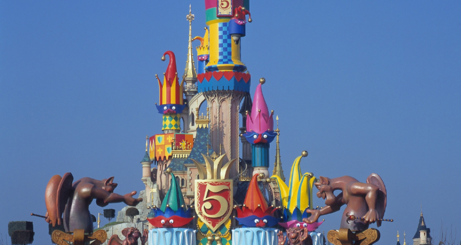 Entrée colorée du château à Disneyland Paris, décorée de grands personnages de dessins animés et d'un emblème « 5 ans », avec des visiteurs qui s'y dirigent sous un ciel bleu limpide.