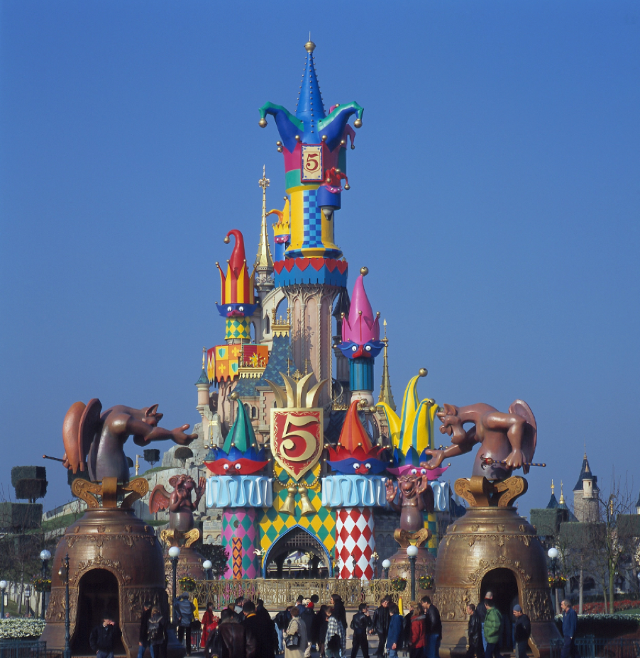 Entrée colorée du château à Disneyland Paris, décorée de grands personnages de dessins animés et d'un emblème « 5 ans », avec des visiteurs qui s'y dirigent sous un ciel bleu limpide.
