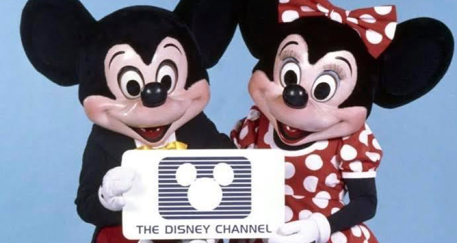 Deux personnes déguisées en Mickey et Minnie Mouse tiennent une pancarte arborant le logo de Disney Channel, célébrant la création de la chaîne le 18 avril 1983, sur un fond bleu uni.