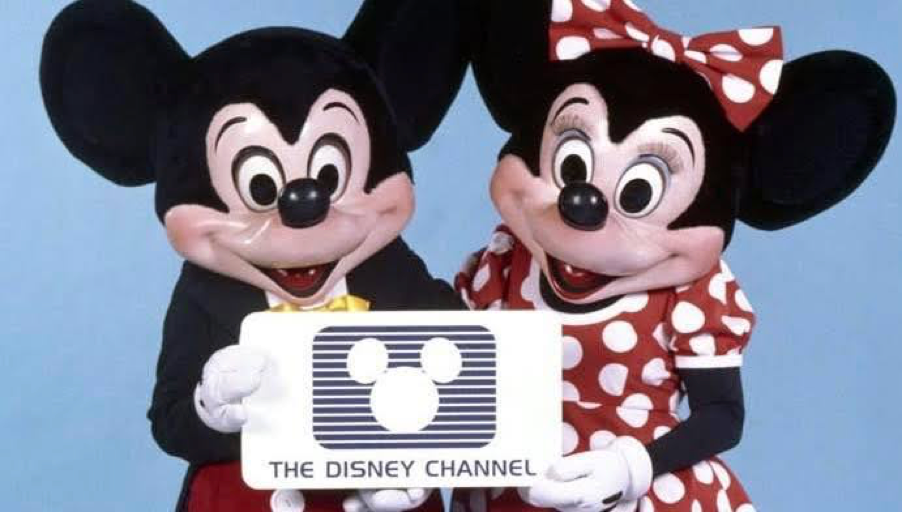 Deux personnes déguisées en Mickey et Minnie Mouse tiennent une pancarte arborant le logo de Disney Channel, célébrant la création de la chaîne le 18 avril 1983, sur un fond bleu uni.