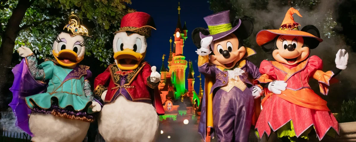 Daisy Duck, Donald Duck, Mickey Mouse et Minnie Mouse, déguisés pour Halloween, posent devant le château illuminé de Disneyland Paris, de nuit.