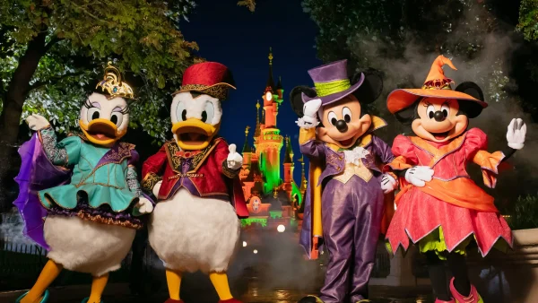 Daisy Duck, Donald Duck, Mickey Mouse et Minnie Mouse, déguisés pour Halloween, posent devant le château illuminé de Disneyland Paris, de nuit.