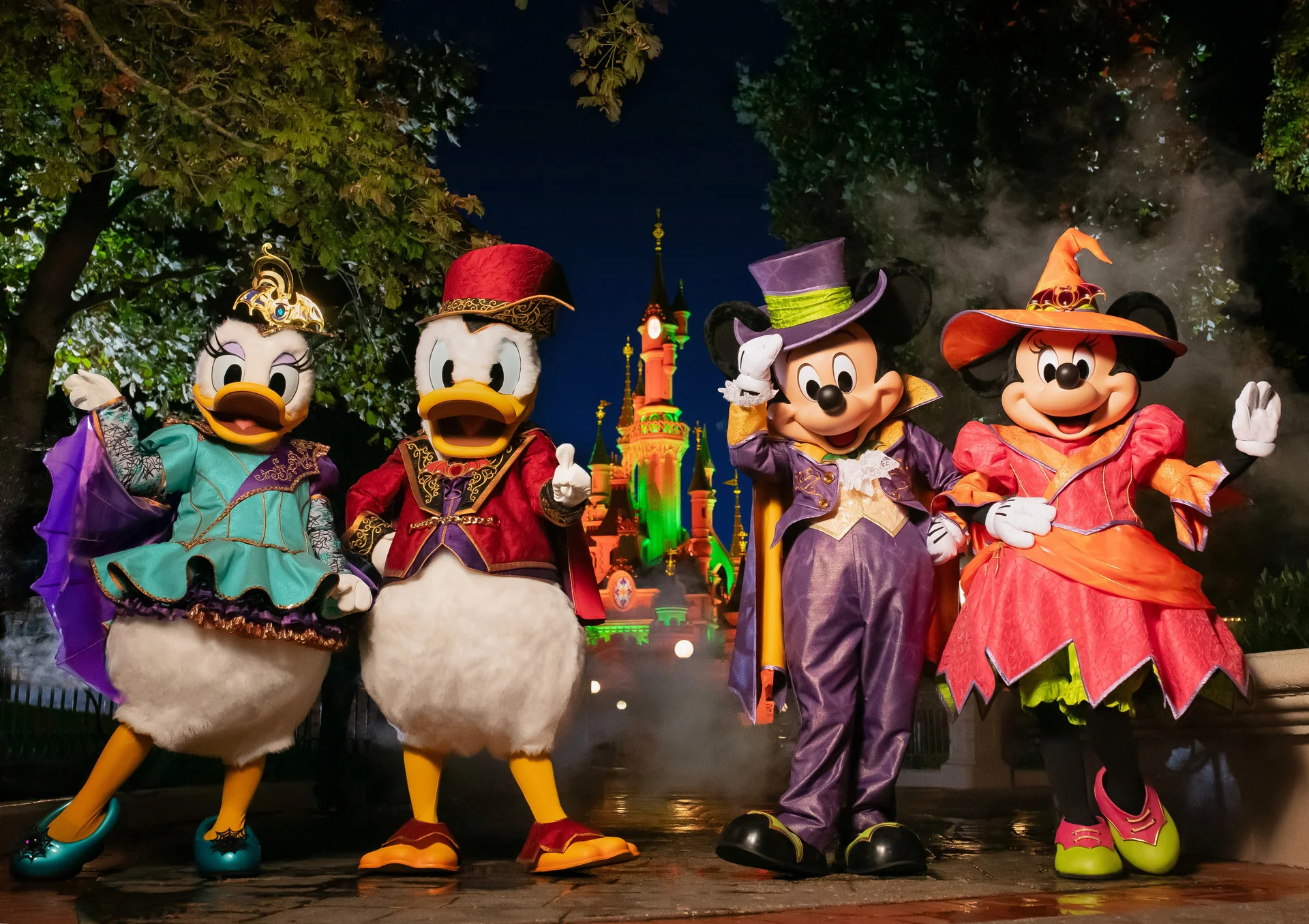 Daisy Duck, Donald Duck, Mickey Mouse et Minnie Mouse, déguisés pour Halloween, posent devant le château illuminé de Disneyland Paris, de nuit.