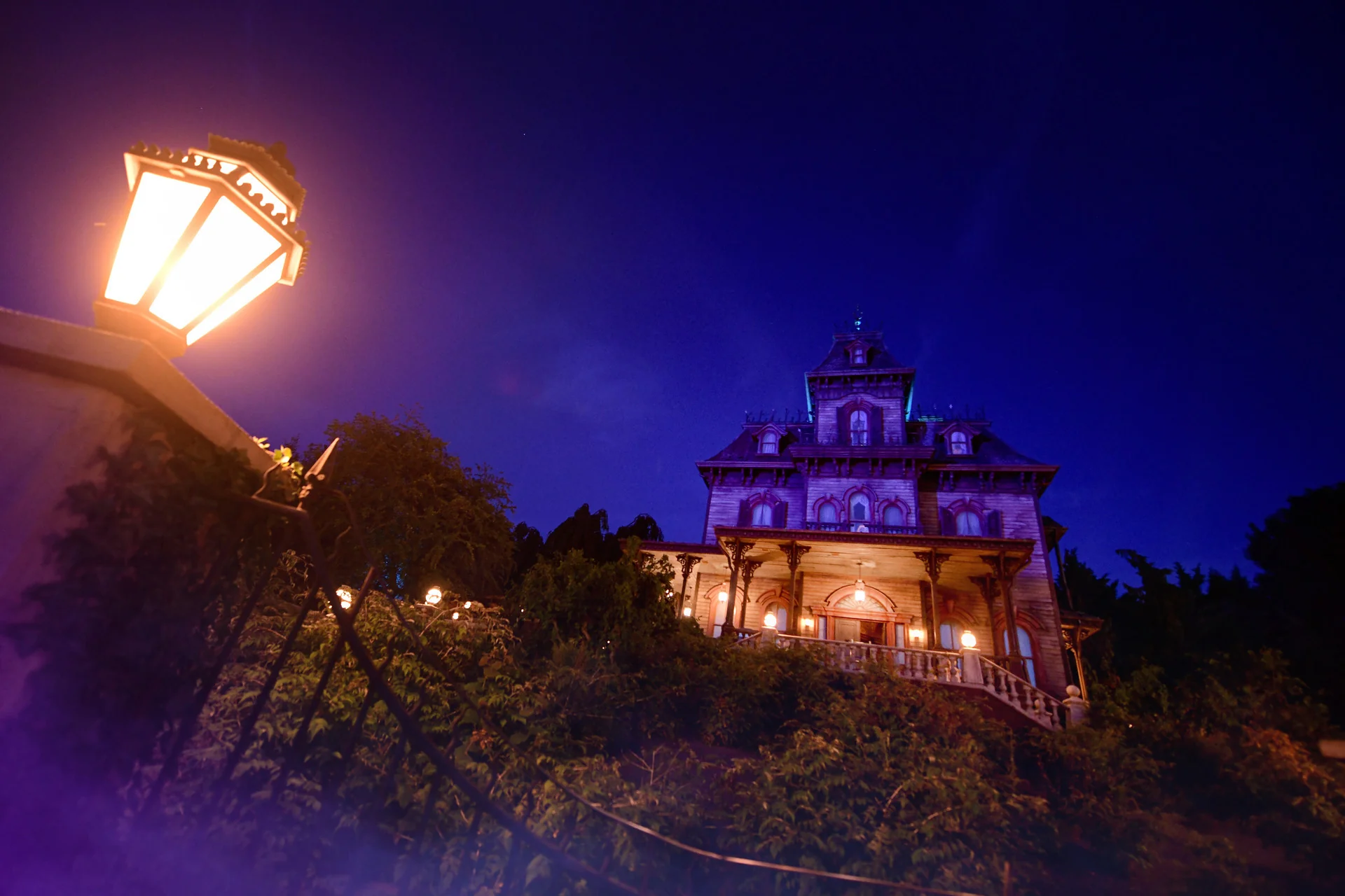 Un manoir sombre de style victorien, aux fenêtres illuminées, se dresse au sommet d'une colline, éclairé par un lampadaire lors du feu d'artifice d'Halloween 2026 à Disneyland Paris.