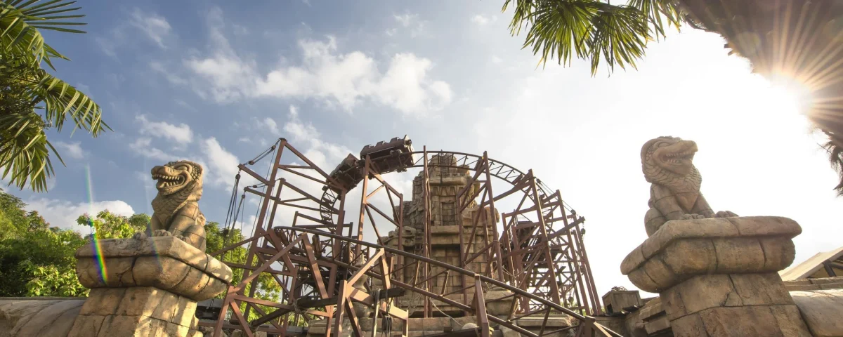 Un wagon de montagnes russes sur le circuit du Temple du Péril passe au-dessus de ruines en pierre flanquées de deux statues de lions sculptées dans la pierre.