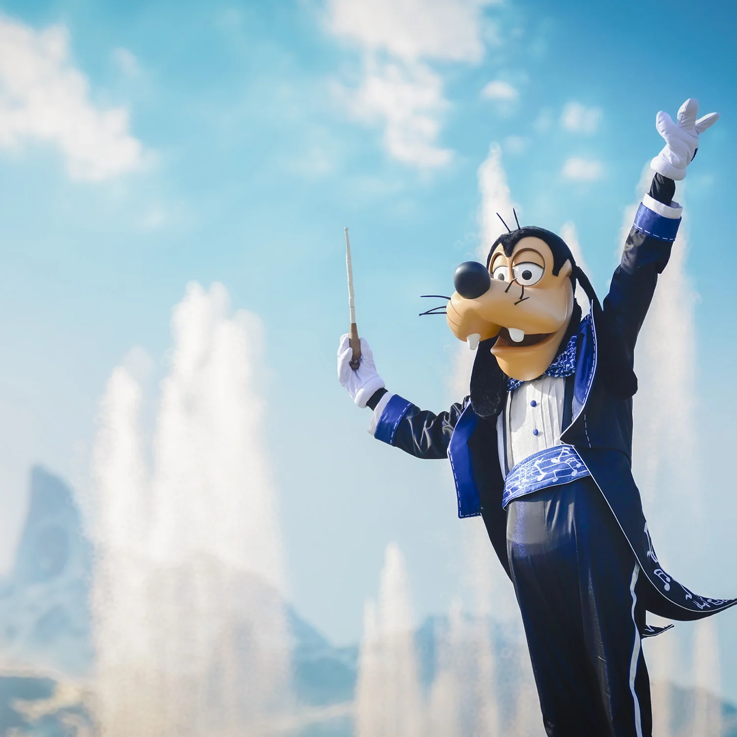 Goofy, déguisé en chef d'orchestre des Dingos, tient une baguette, le bras levé, devant une fontaine.