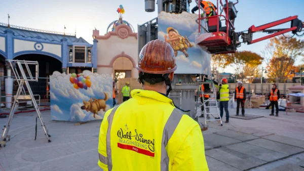 Un Imagineer de Disney, vêtu d'un gilet de sécurité et d'un casque de chantier, observe des ouvriers en train d'installer une structure sur le thème des « Wilderness Explorers » du film « Là-haut » dans un parc d'attractions.