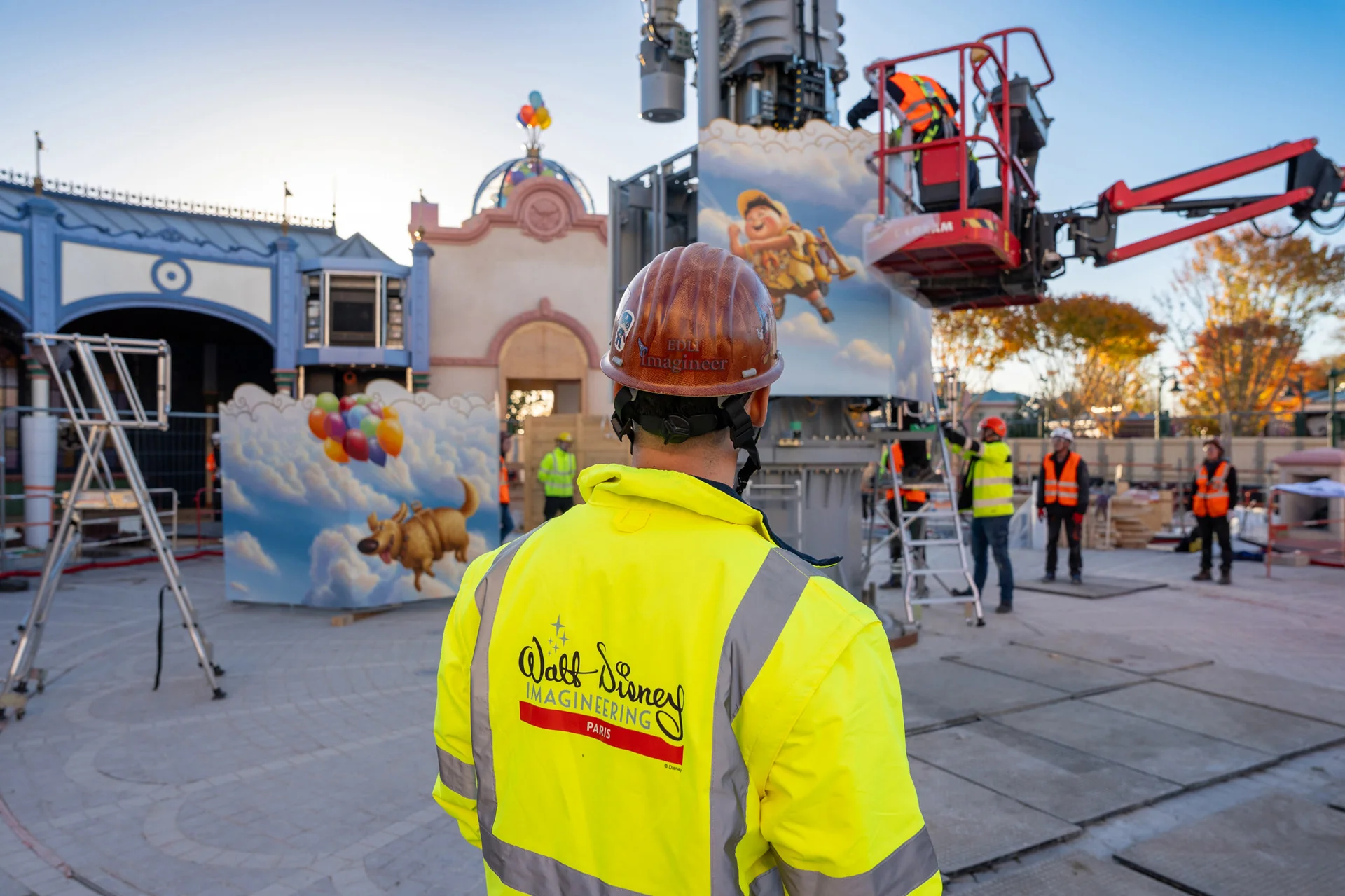 Un Imagineer de Disney, vêtu d'un gilet de sécurité et d'un casque de chantier, observe des ouvriers en train d'installer une structure sur le thème des « Wilderness Explorers » du film « Là-haut » dans un parc d'attractions.